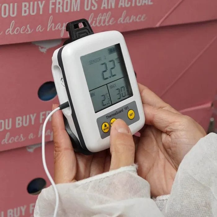 une personne tenant un Thermomètre Enregistreur de surveillance de la température du réfrigérateur ThermaGuard de Thermometre.fr devant une boîte rose.